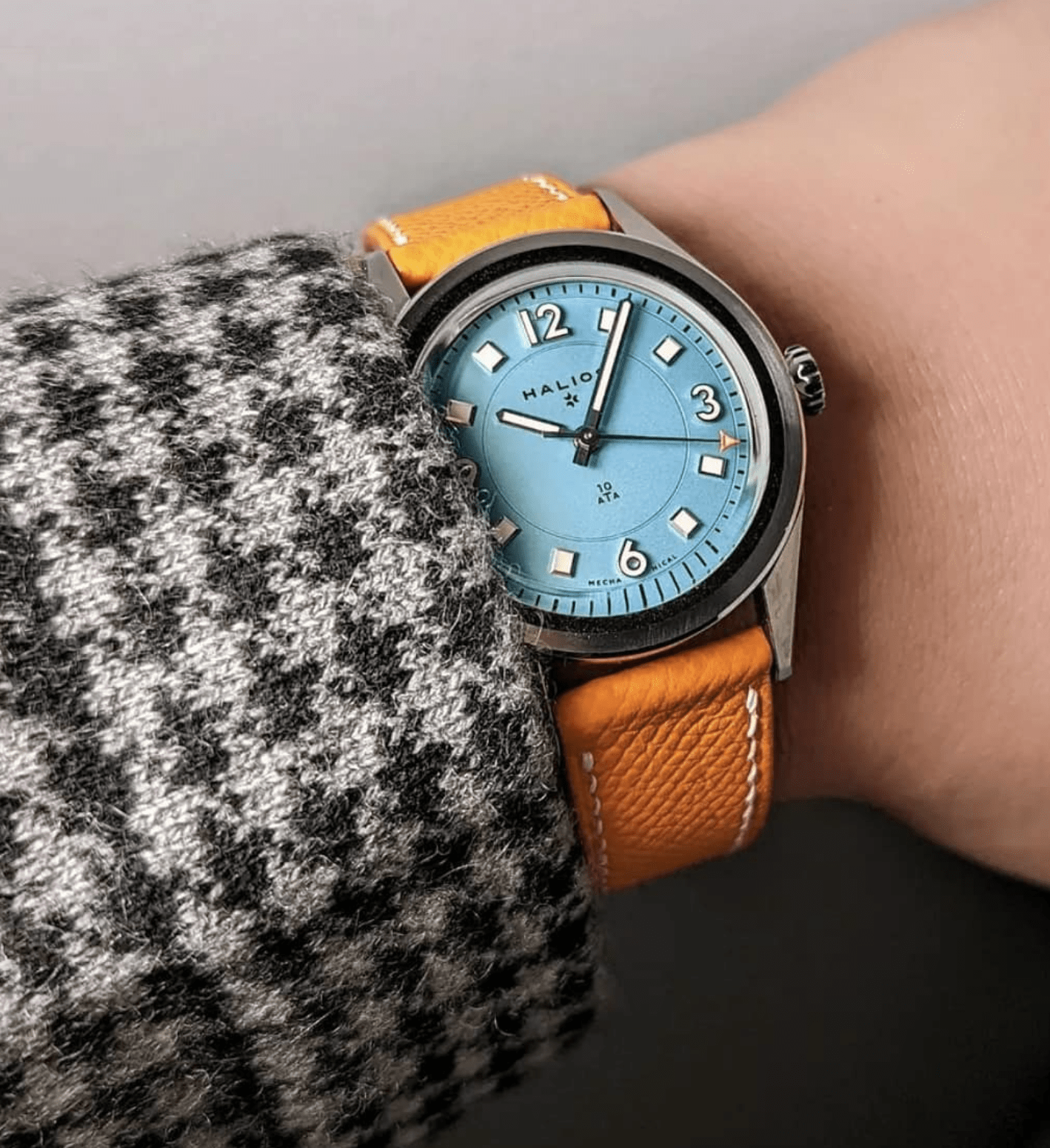 Watch with a blue face and orange strap on a wrist, wearing a checkered coat.