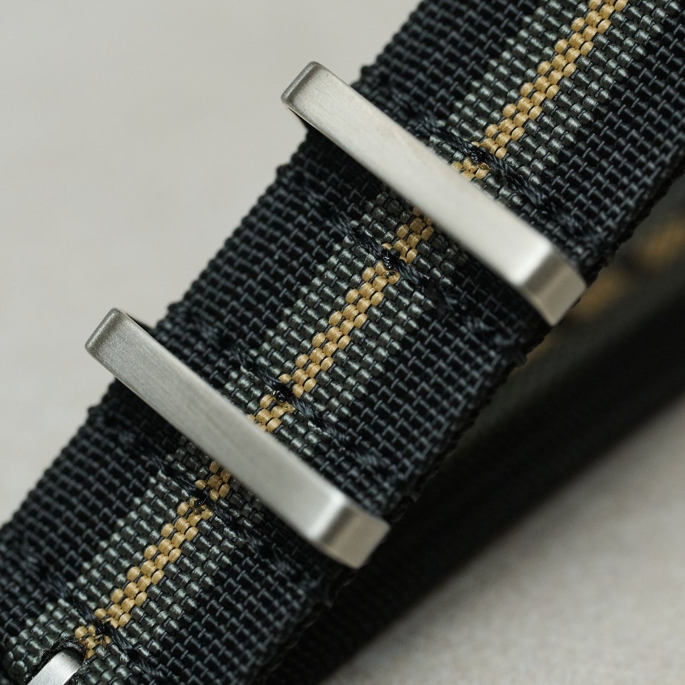 Close-up of a woven watch strap in black and gray with a thin gold stripe, featuring brushed silver metal buckle hardware.