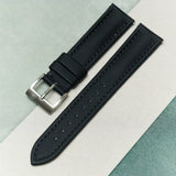A black textured watch strap with a silver-toned metal buckle, laid diagonally on a split background of light gray and mint green.