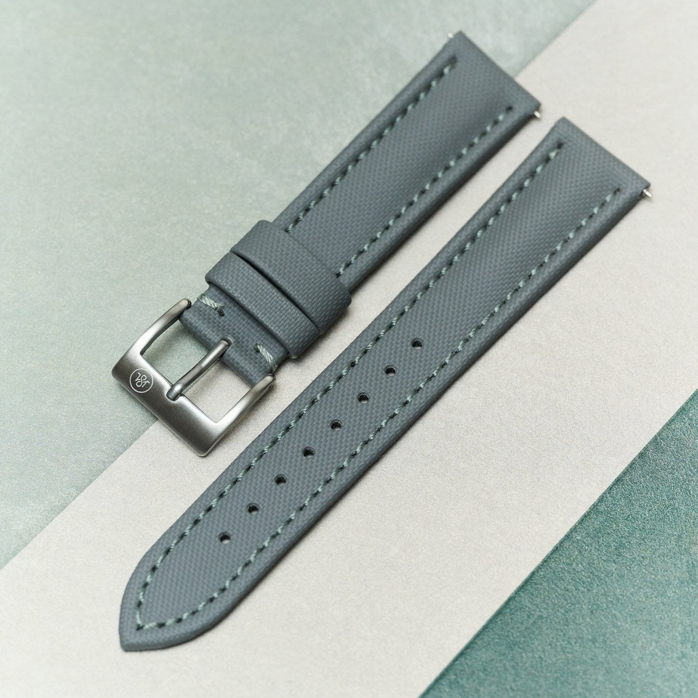 A gray textured leather watch strap with a silver-toned metal buckle, laid diagonally on a split beige and sage green background.