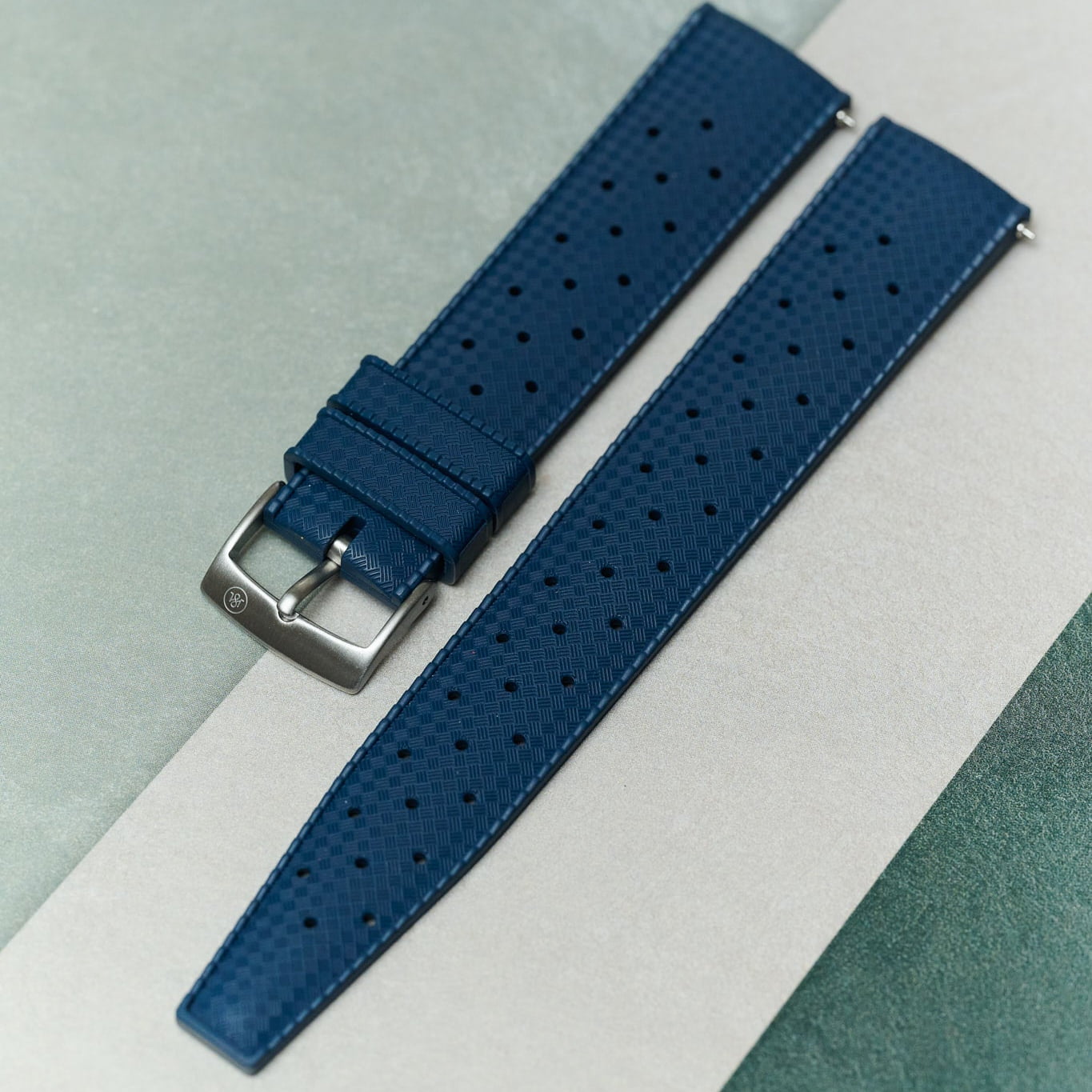 A blue perforated leather watch strap with a silver-toned metal buckle, laid diagonally on a split light gray and textured teal background.