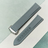 A gray textured leather watch strap with a silver-toned metal buckle, laid diagonally on a split background of light gray and teal-green textured paper.