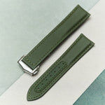 An olive green textured watch strap with a silver-toned metal buckle, laid diagonally on a split background of light gray and teal.