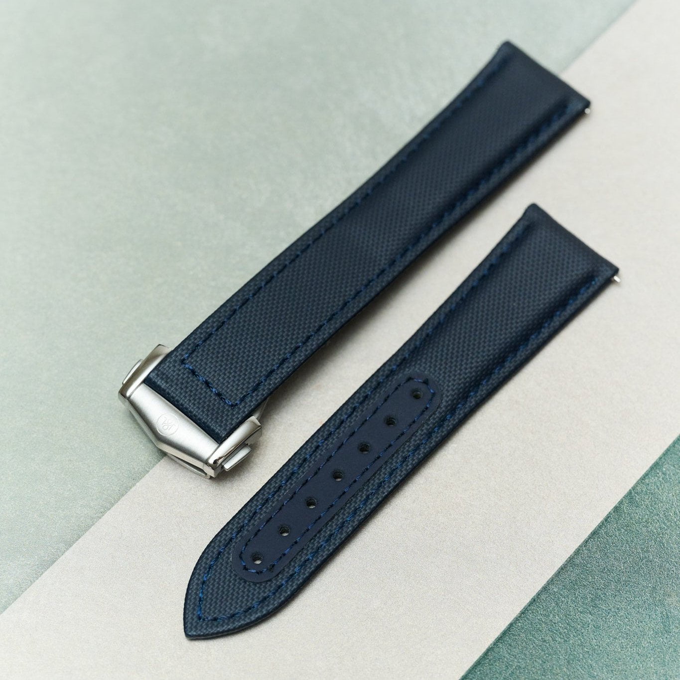 A dark navy blue textured leather watch strap with a silver-toned metal buckle, laid diagonally on a split white and light green background.