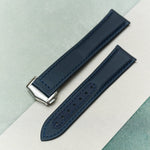 A dark navy blue textured leather watch strap with a silver-toned metal buckle, laid diagonally on a split white and light green background.