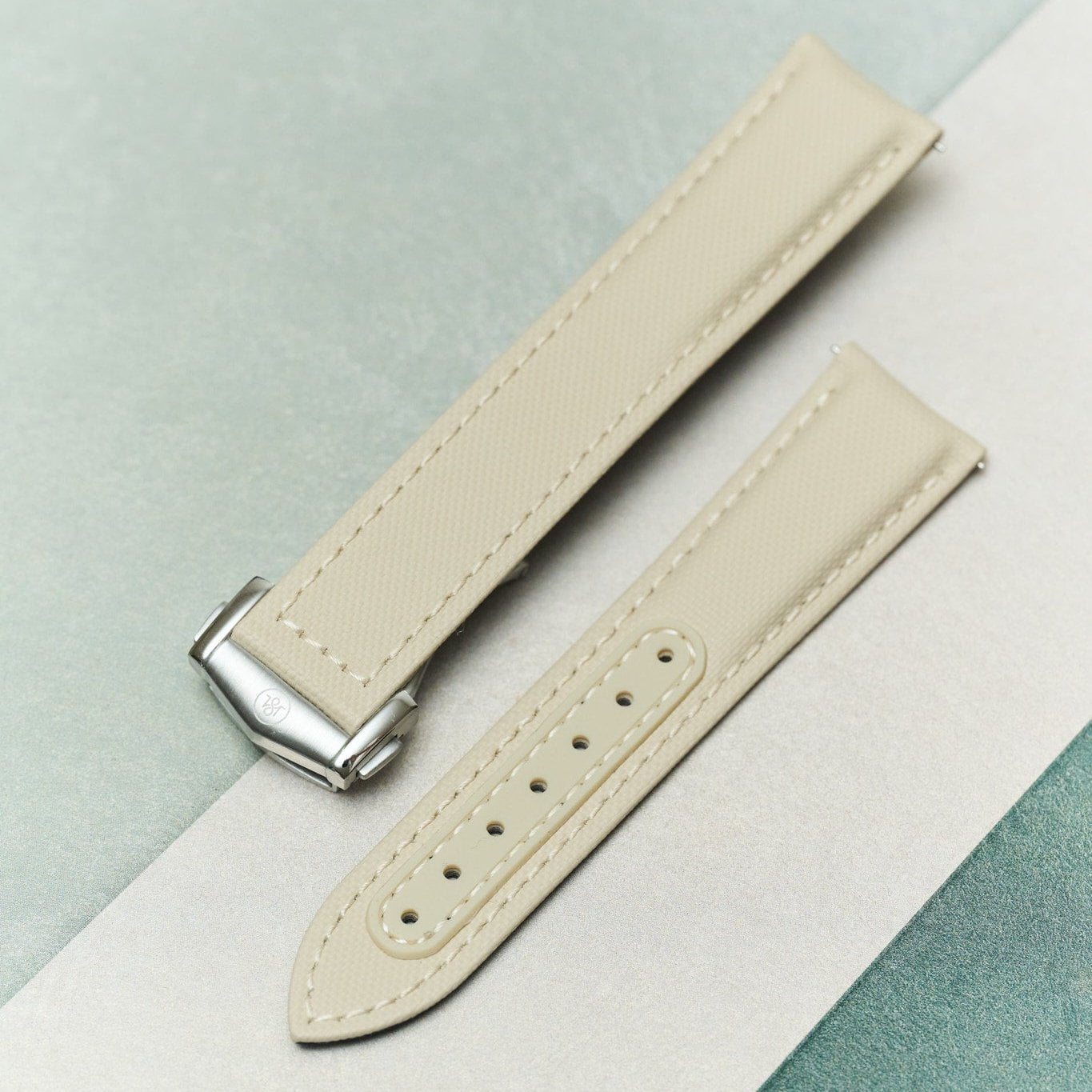 A light beige leather watch strap with a silver-toned metal buckle, laid diagonally on a split white and mint green background.