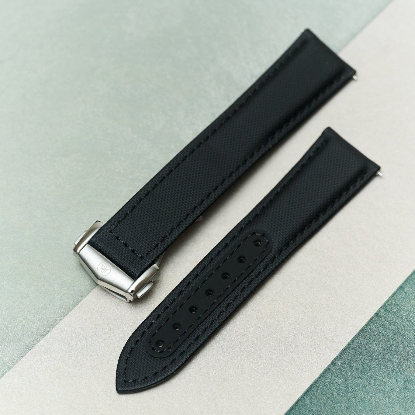 A black textured watch strap with a silver-toned metal buckle, laid diagonally on a split light gray and mint green background.
