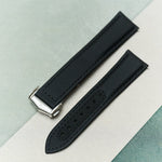 A black textured watch strap with a silver-toned metal buckle, laid diagonally on a split light gray and mint green background.