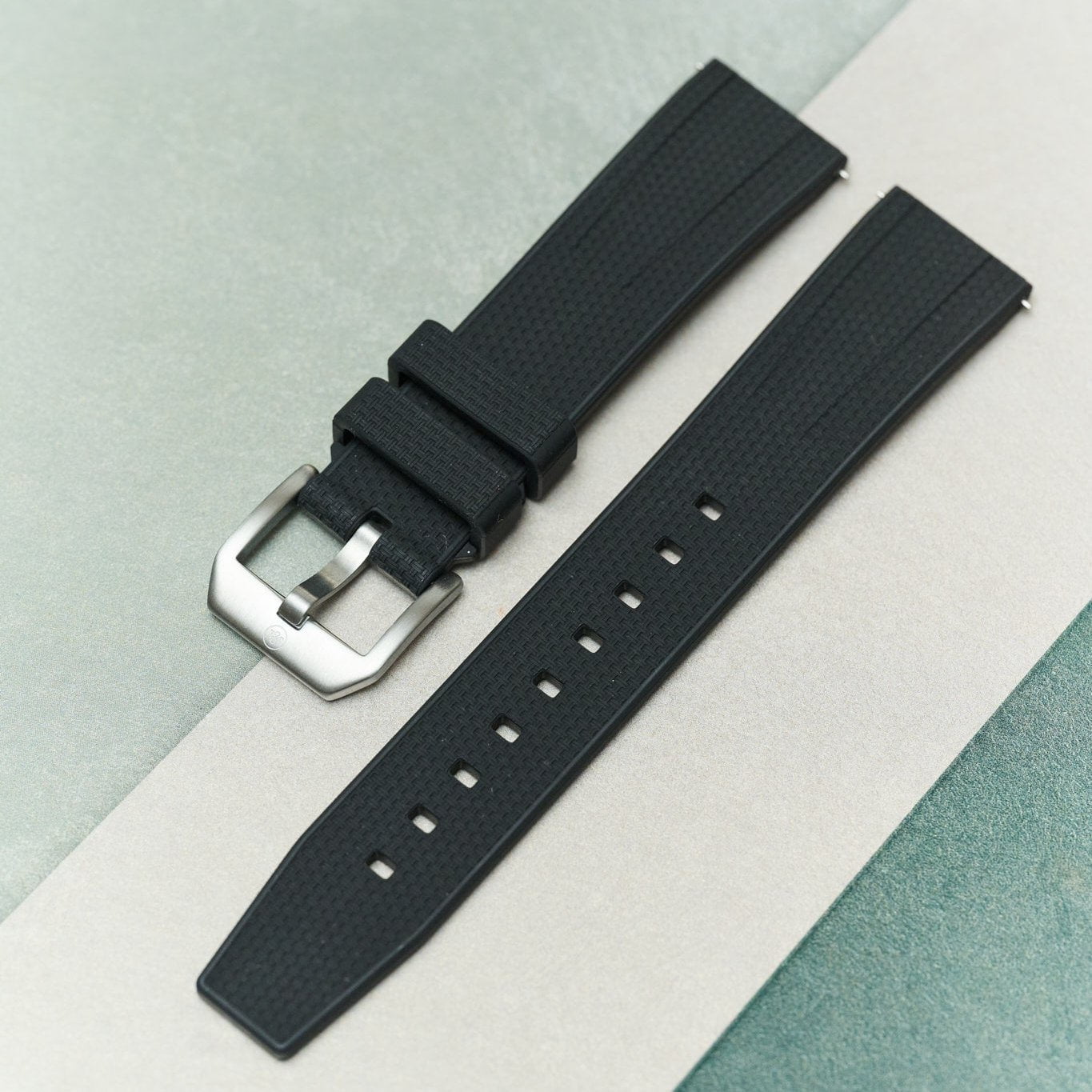A black textured rubber watch strap with a silver-toned metal buckle, laid diagonally on a split light gray and teal background.