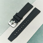A black textured rubber watch strap with a silver-toned metal buckle, laid diagonally on a split light gray and teal background.