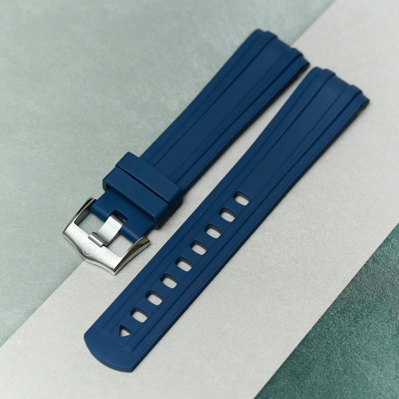 A dark blue silicone watch strap with a silver metal buckle, laid diagonally on a split light gray and textured teal background.
