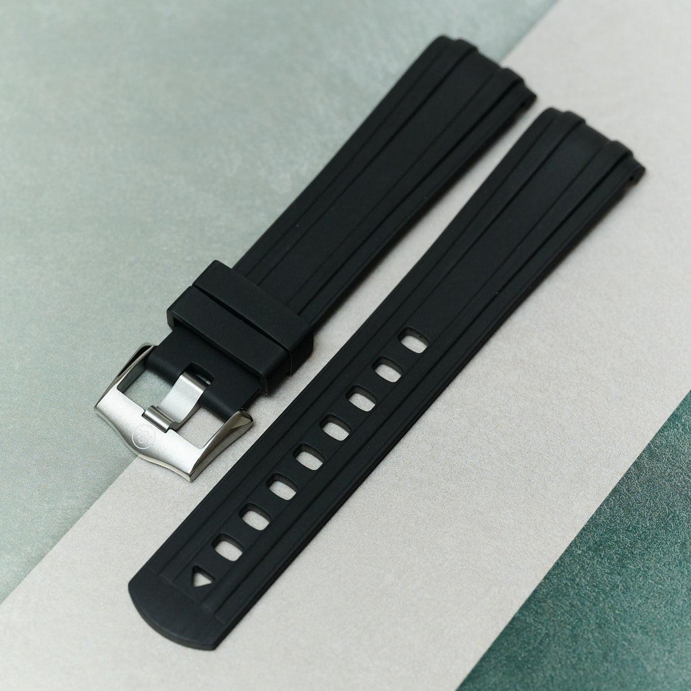 A black silicone watch strap with a silver metal buckle, laid diagonally on a split light gray and green textured background.