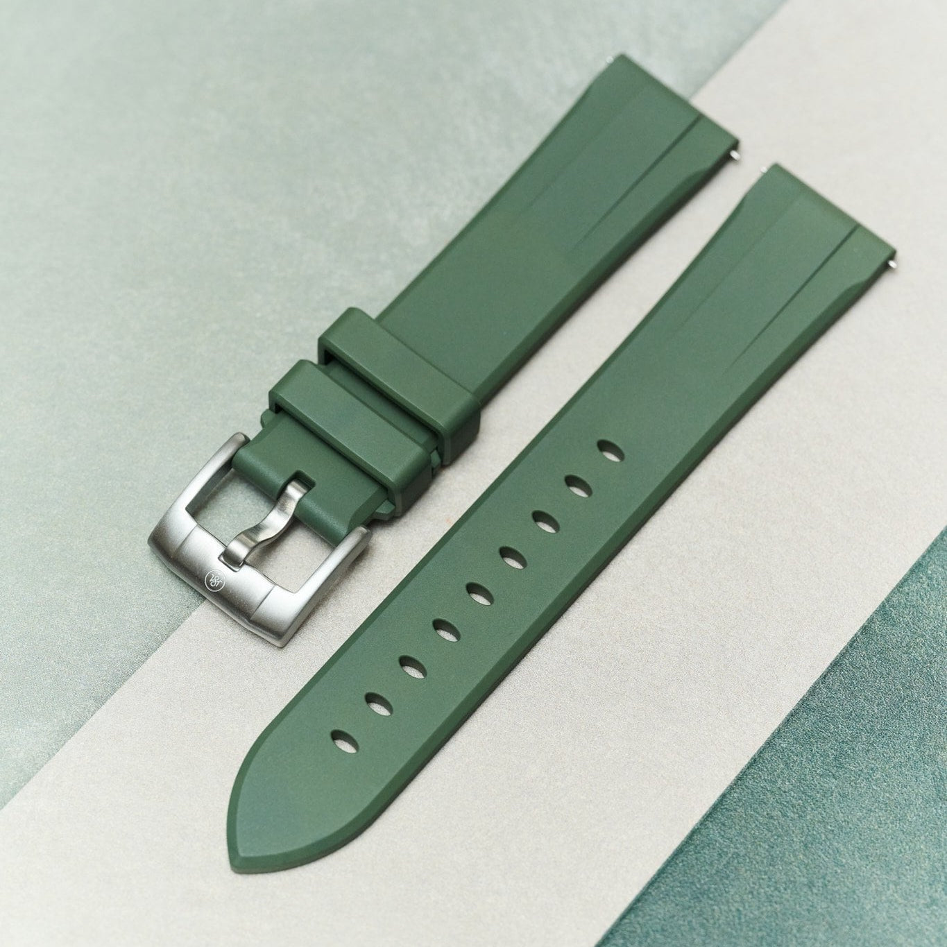 A green silicone watch strap with a silver metal buckle, laid out diagonally on a split light gray and teal textured background.