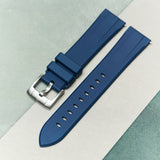 A dark blue silicone watch strap with a silver-toned metal buckle, laid diagonally on a split background of light gray and pale green.