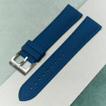 A textured blue watch strap with a silver metal buckle, laid diagonally on a split background of sage green and off-white.