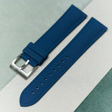 A textured blue watch strap with a silver metal buckle, laid diagonally on a split background of sage green and off-white.