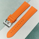 An orange textured silicone watch strap with a silver metal buckle, laid diagonally on a split white and sage green background.