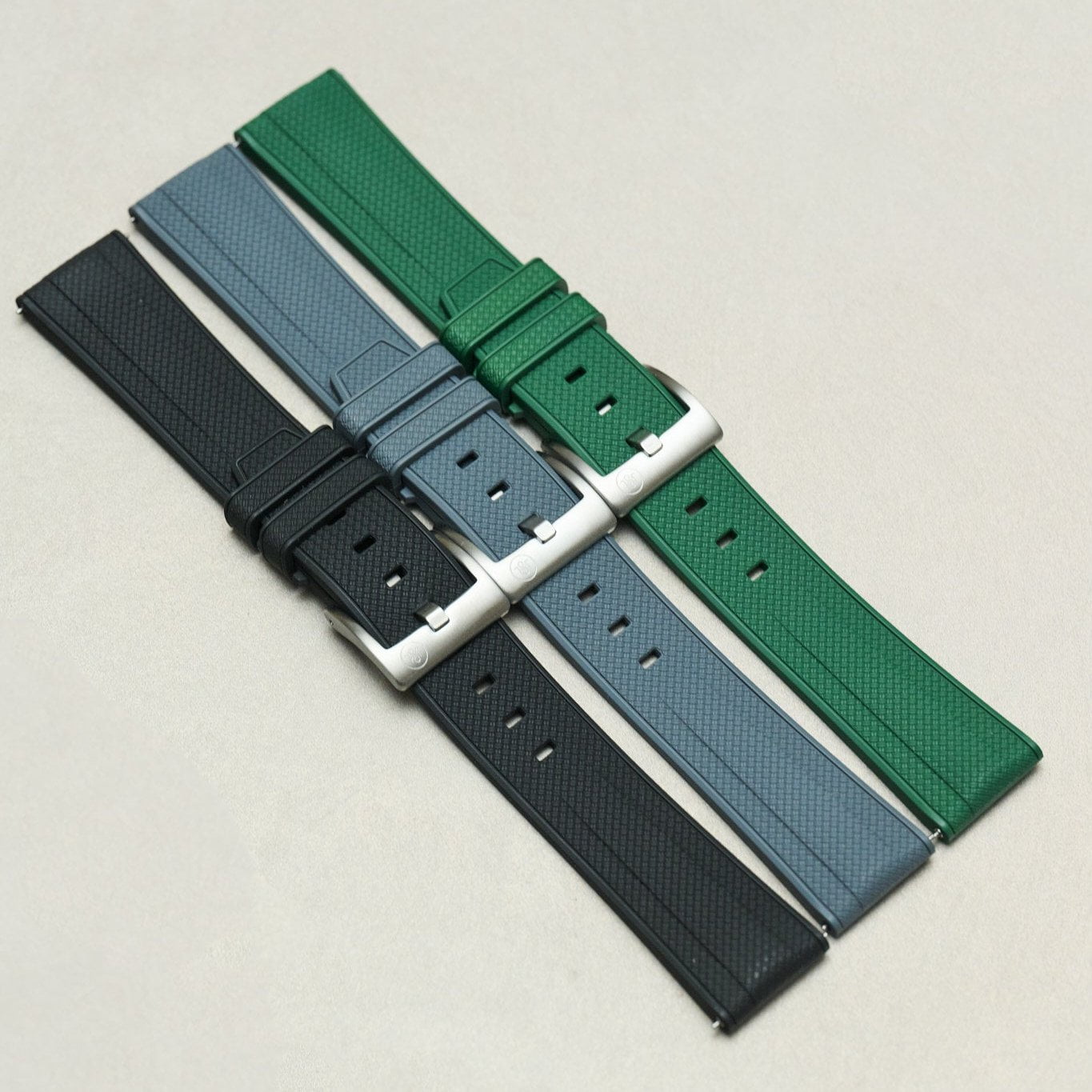 Three textured watch straps in black, slate blue, and forest green, each with a silver-toned metal buckle, arranged diagonally on a light beige background.