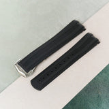 A black rubber watch strap with a silver-toned metal buckle, laid diagonally on a split background of light gray and dark green textured surfaces.